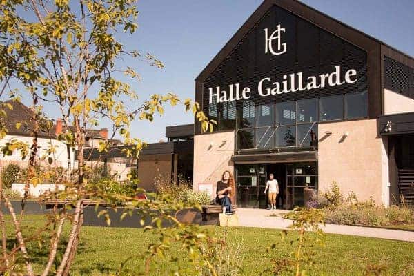 Vue sur l'extérieur de la Halle gaillarde. Un couple est assis sur un banc devant l'entrée