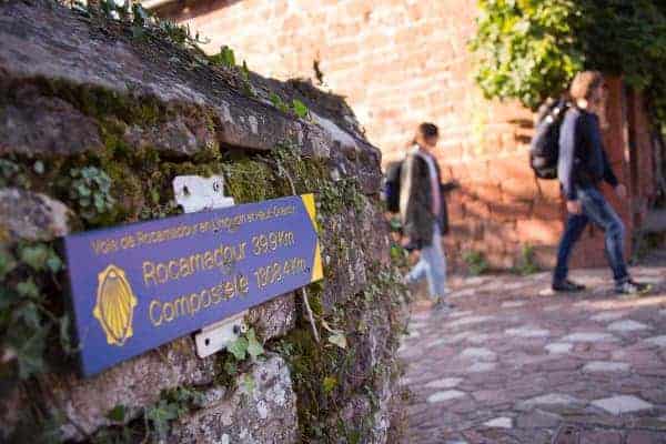 Vue sur un panneau indiquant le chemin de Compostelle avec un couple de marcheurs à l'arrière-plan