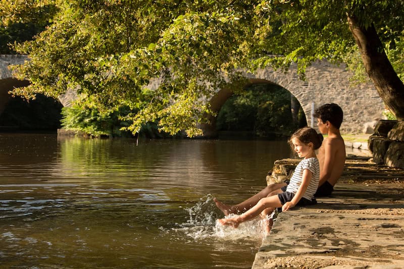 Jeunes enfants assis sur une margelle devant le vieux pont du Saillant et faisant des éclaboussures avec leur pieds dans l'eau de la Vézère
